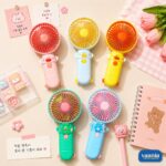 Five colorful CuteSpin Hand Fans in pink, blue, yellow, and green arranged on a desk with cute stationery.