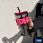 A hand holding a smoked grey Hoho Glass Sipper with a bright pink silicone bubble grip and matching lid.