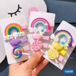 A hand holding three cards of Rainbow Candy Hair Clips featuring colorful rainbows, clouds, and swirl lollipops.