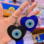 Two heart-shaped Evil Eye Keychains in black and blue being held by a hand showing crystal details.