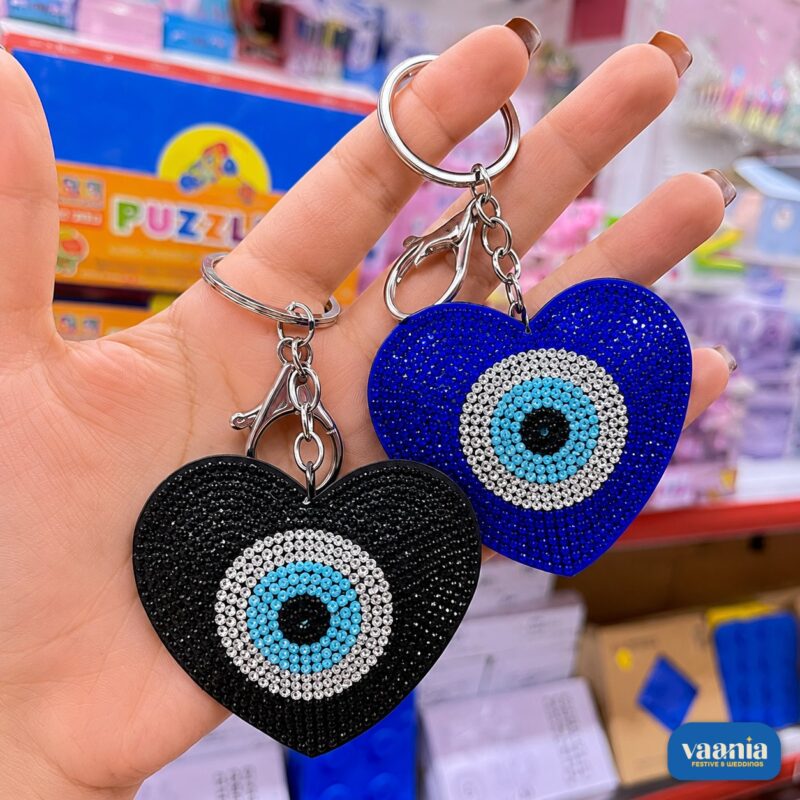 Two heart-shaped Evil Eye Keychains in black and blue being held by a hand showing crystal details.