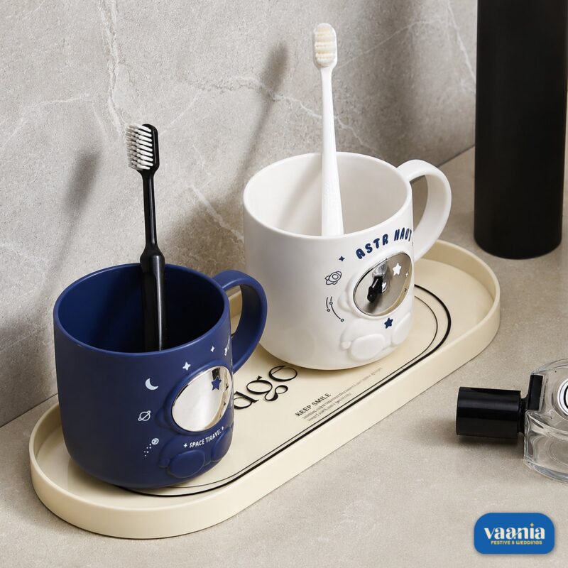 Blue and white Astronaut Buddy Mugs holding toothbrushes on a cream tray in a bathroom setting.