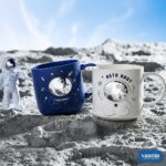 Blue and white Astronaut Buddy Mugs holding toothbrushes on a cream tray in a bathroom setting. - view 2