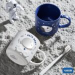 Blue and white Astronaut Buddy Mugs holding toothbrushes on a cream tray in a bathroom setting. - view 3