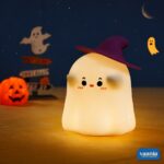 A glowing Ghostess Silicone Night Lamp wearing a purple witch hat sitting on a nightstand. - view 3