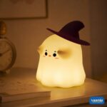 A glowing Ghostess Silicone Night Lamp wearing a purple witch hat sitting on a nightstand.