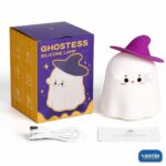 A glowing Ghostess Silicone Night Lamp wearing a purple witch hat sitting on a nightstand. - view 6
