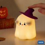 A glowing Ghostess Silicone Night Lamp wearing a purple witch hat sitting on a nightstand. - view 4