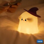 A glowing Ghostess Silicone Night Lamp wearing a purple witch hat sitting on a nightstand. - view 5
