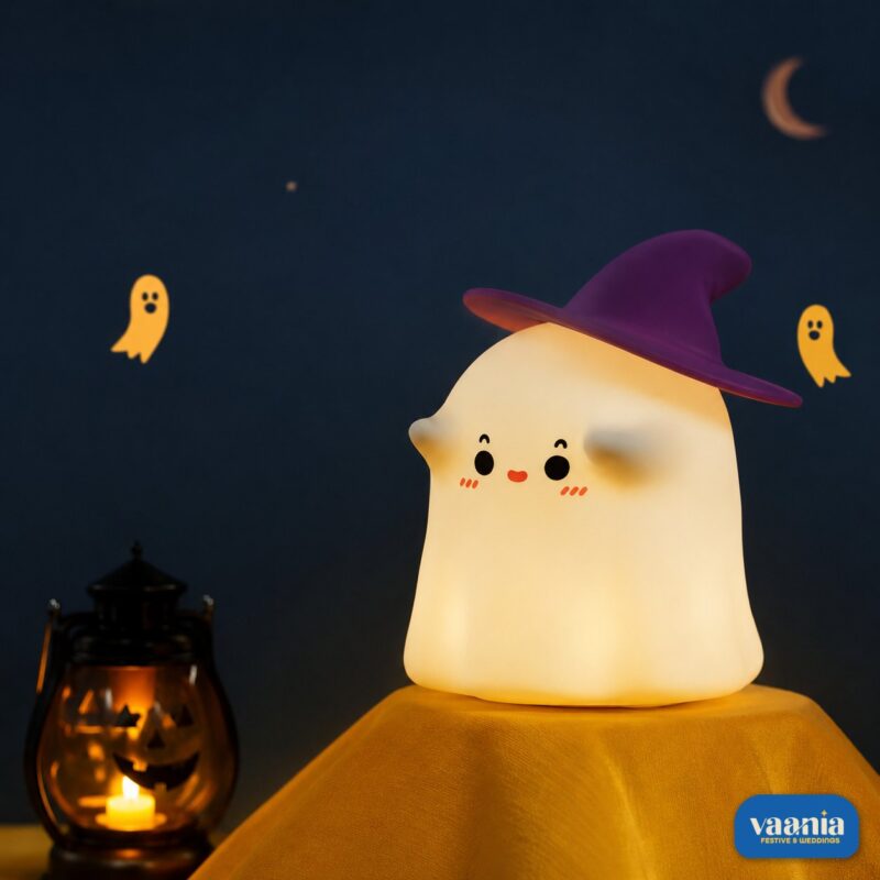 A glowing Ghostess Silicone Night Lamp wearing a purple witch hat sitting on a nightstand. - view 2
