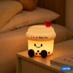 A glowing Boba Tea Silicone Night Lamp with a smiley face and heart topper sitting next to two macarons. - view 5