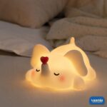 A glowing Loving Elephant Silicone Night Lamp with a red heart on its trunk sitting on a bed.