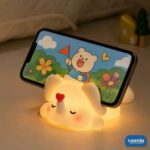 A glowing Loving Elephant Silicone Night Lamp with a red heart on its trunk sitting on a bed. - view 3
