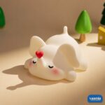 A glowing Loving Elephant Silicone Night Lamp with a red heart on its trunk sitting on a bed. - view 4