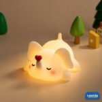 A glowing Loving Elephant Silicone Night Lamp with a red heart on its trunk sitting on a bed. - view 5