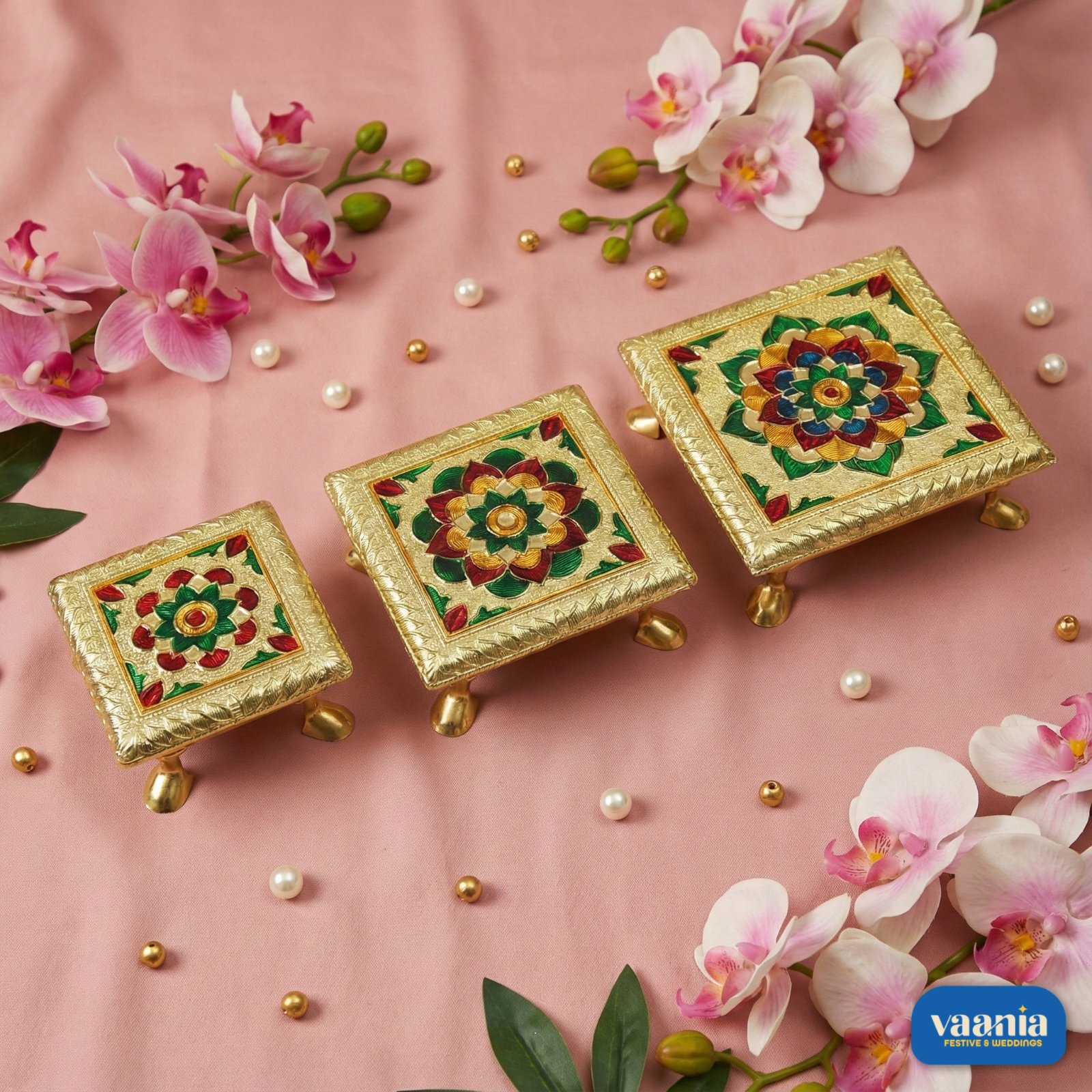 Vaania Designer Metal Leg Chowki Set Three golden Metal Leg Chowkis with colorful floral patterns and curved brass feet on a pink backdrop.