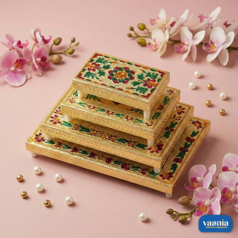 A stack of four golden nested Rectangular Chowkis with floral paintings surrounded by pink orchids and pearls.