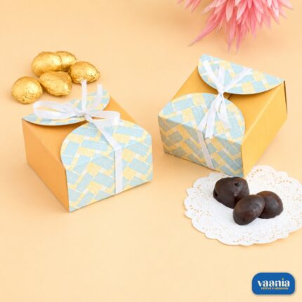 Two yellow Multi Purpose Boxes with blue patterned lids and white ribbons next to gold and dark chocolates.