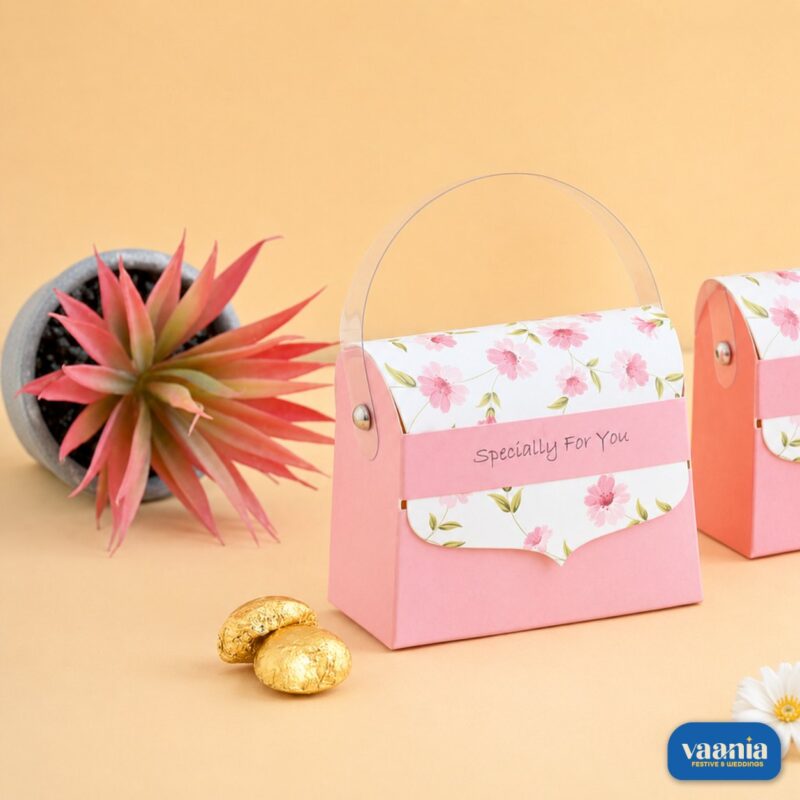 A pink Mini Hand Bag Box with floral prints and a transparent handle sitting next to gold chocolates and a small plant.