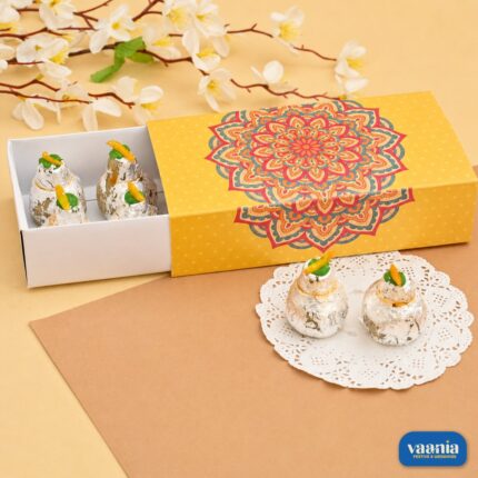 A yellow Sliding Box with a colorful mandala design containing silver-coated sweets on a beige surface.