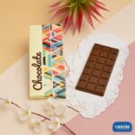 A dark milk chocolate bar resting on a white lace doily next to a colorful geometric Chocolate Bar Box.