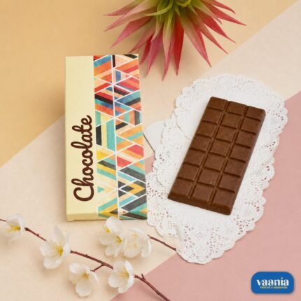 A dark milk chocolate bar resting on a white lace doily next to a colorful geometric Chocolate Bar Box.