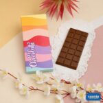 A milk chocolate bar next to a colorful wavy patterned Chocolate Bar Box with floral accents.