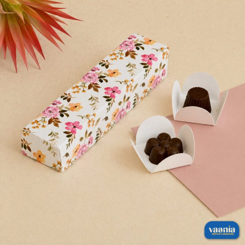 A white floral pattern 4 cavity box next to two gourmet chocolates in white paper liners.