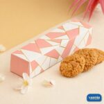A rectangular Multi Purpose Box with pink and white geometric patterns next to three golden-brown oat cookies and small white flowers.