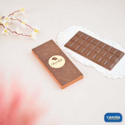 A smooth rectangular milk chocolate bar next to a decorative brown Chocolate Bar Box on a white lace doily.
