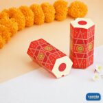 Two red hexagonal Dhol Boxes with traditional patterns next to yellow marigold flowers and white blossoms.