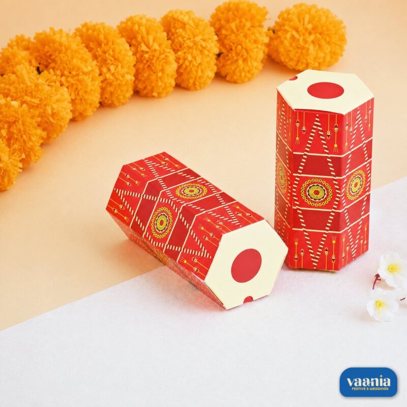 Two red hexagonal Dhol Boxes with traditional patterns next to yellow marigold flowers and white blossoms.