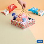 A hand lifting a colorful geometric patterned Hamper Tray filled with drinks and snacks by its golden rope handles.