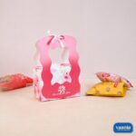 A pink floral Hamper Box with a white ribbon and wavy handle sitting next to assorted snack pouches.