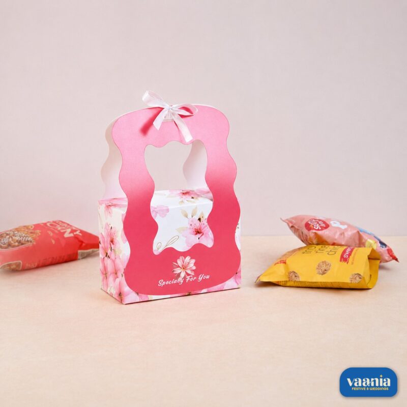 A pink floral Hamper Box with a white ribbon and wavy handle sitting next to assorted snack pouches.