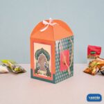 An orange and teal Hamper Box with peacock designs, ribbon top, and rope handles surrounded by snack packets.