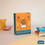 A decorative orange and teal Gift Carry Bag featuring a white cow motif, surrounded by snacks on a pastel background.