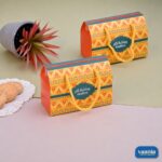 Two orange and yellow patterned Vaania hamper boxes with yellow rope handles alongside cookies and a potted plant.