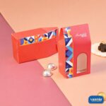 Two red Window Candy Boxes with geometric patterns and silver chocolates on a pink and peach background.