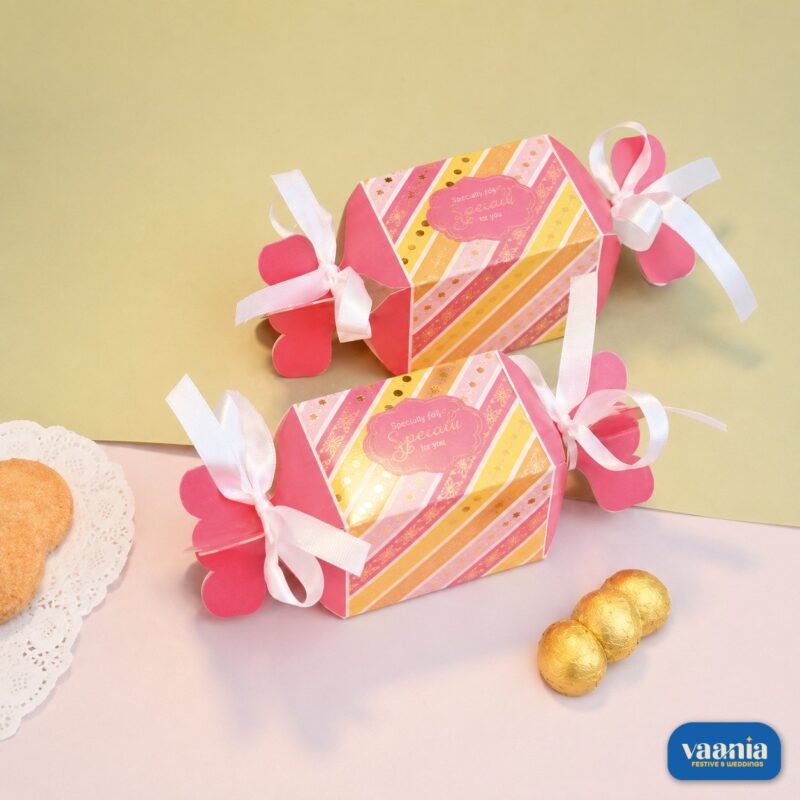 Two pink and gold striped Toffee Shape Boxes with white ribbons beside gold-wrapped chocolates.