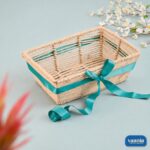 A hand-woven rectangular jute hamper basket featuring an open-slat design and a decorative teal satin ribbon bow.