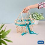 A hand holding a natural jute Handle Hamper Basket with a teal ribbon bow on a table near a houseplant.