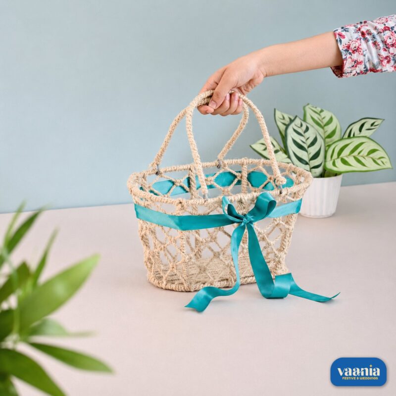 A hand holding a natural jute Handle Hamper Basket with a teal ribbon bow on a table near a houseplant.