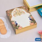 A luxury gold Sliding Box featuring vibrant peacock and floral artwork on the lid next to cookies.