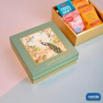 A luxury Top-Bottom Hamper with a peacock design lid next to an open gold box filled with colorful tea packets.