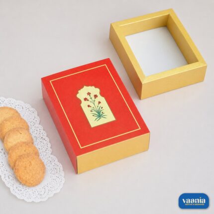 A red sliding box with floral art next to a gold tray and cookies on a white doily.