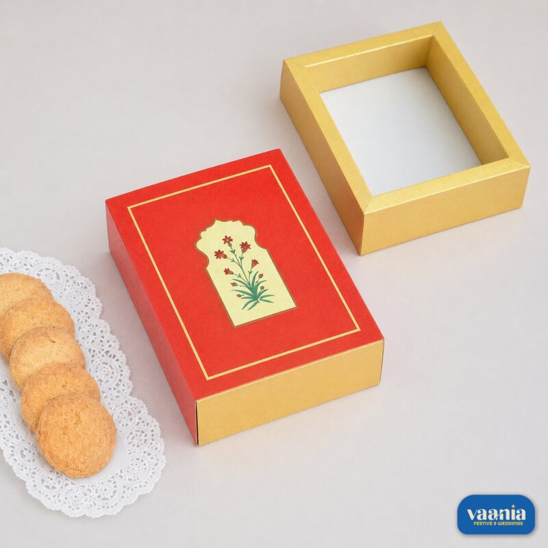 A red sliding box with floral art next to a gold tray and cookies on a white doily.