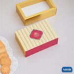 A decorative beige and pink striped Sliding Box next to an open gold tray and fresh cookies.