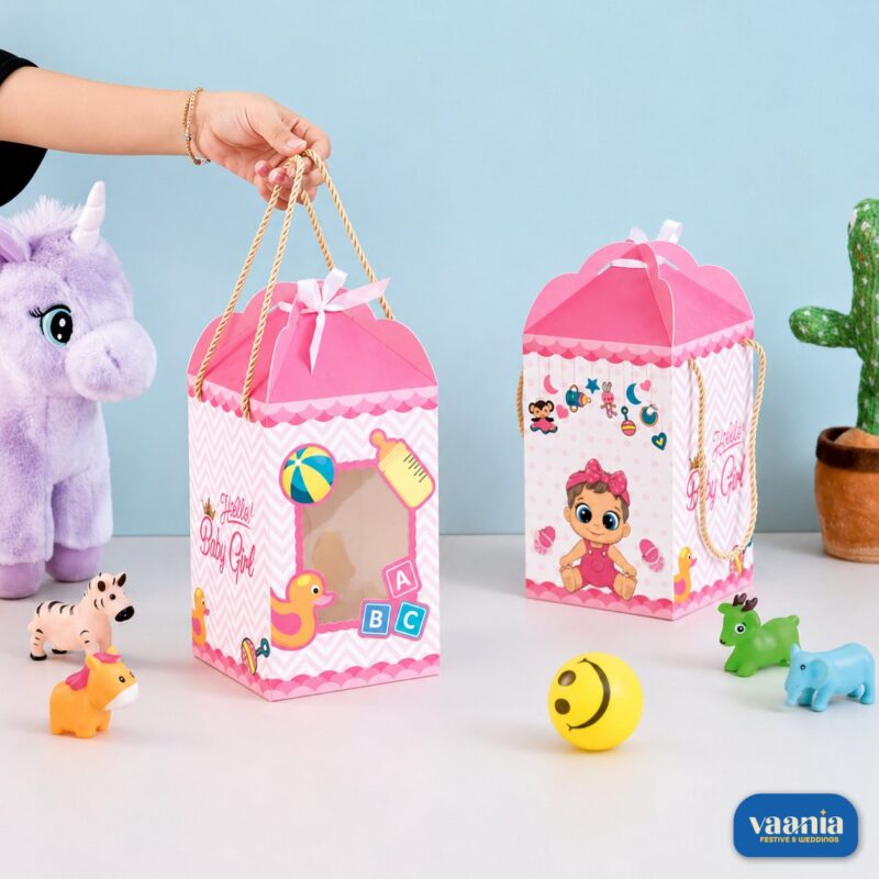 A hand holding a pink Hamper Box (Baby Girl) featuring a window and baby illustrations next to a purple plush toy.