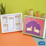 Traditional Pichwai cow design Book Shape Hamper Box with five gold-capped PET jars on an orange and green surface.